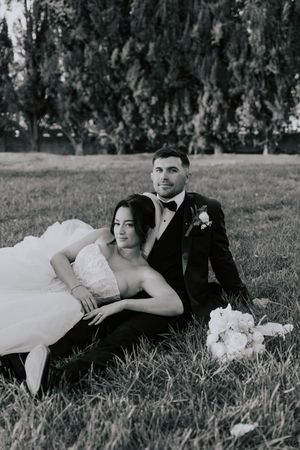 Black and white photo of newlyweds relaxing on grass, bride leaning on groom in tuxedo.