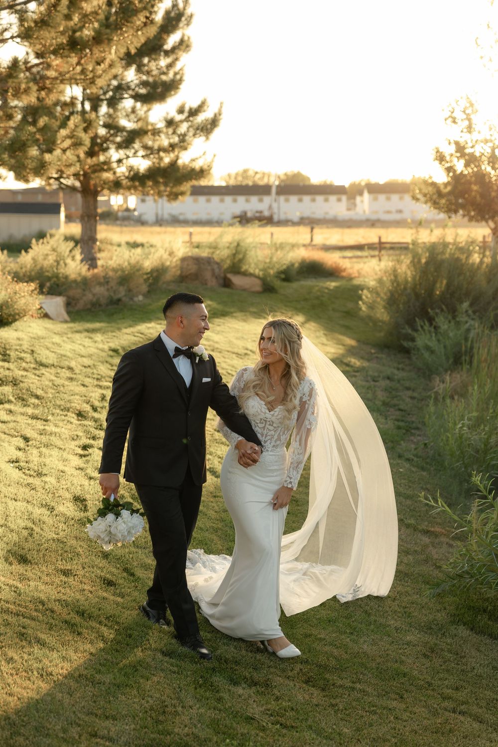 Bride and groom walking hand in hand at sunset with veil flowing in the breeze.