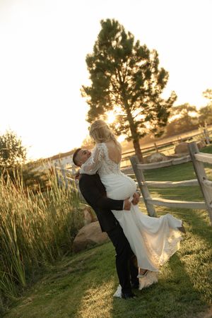 Groom lifting bride in white lace dress at golden hour near wooden fence and tall trees.