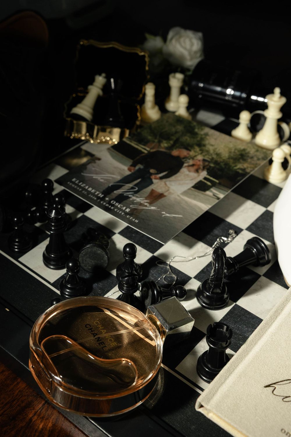 Chess board with scattered black and white pieces, perfume bottle, lighter, and elegant stationery in moody lighting.