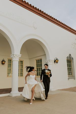 Bride and groom walking hand in hand outside white Spanish colonial building with arched columns.