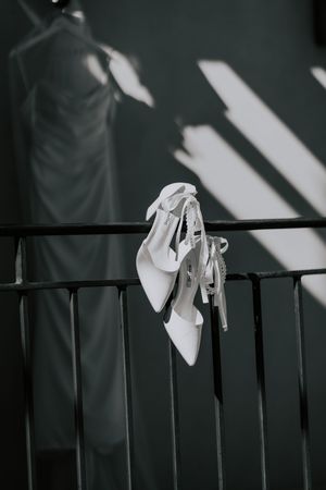 Black and white photo of white bridal heels hanging on iron railing with dress in background.