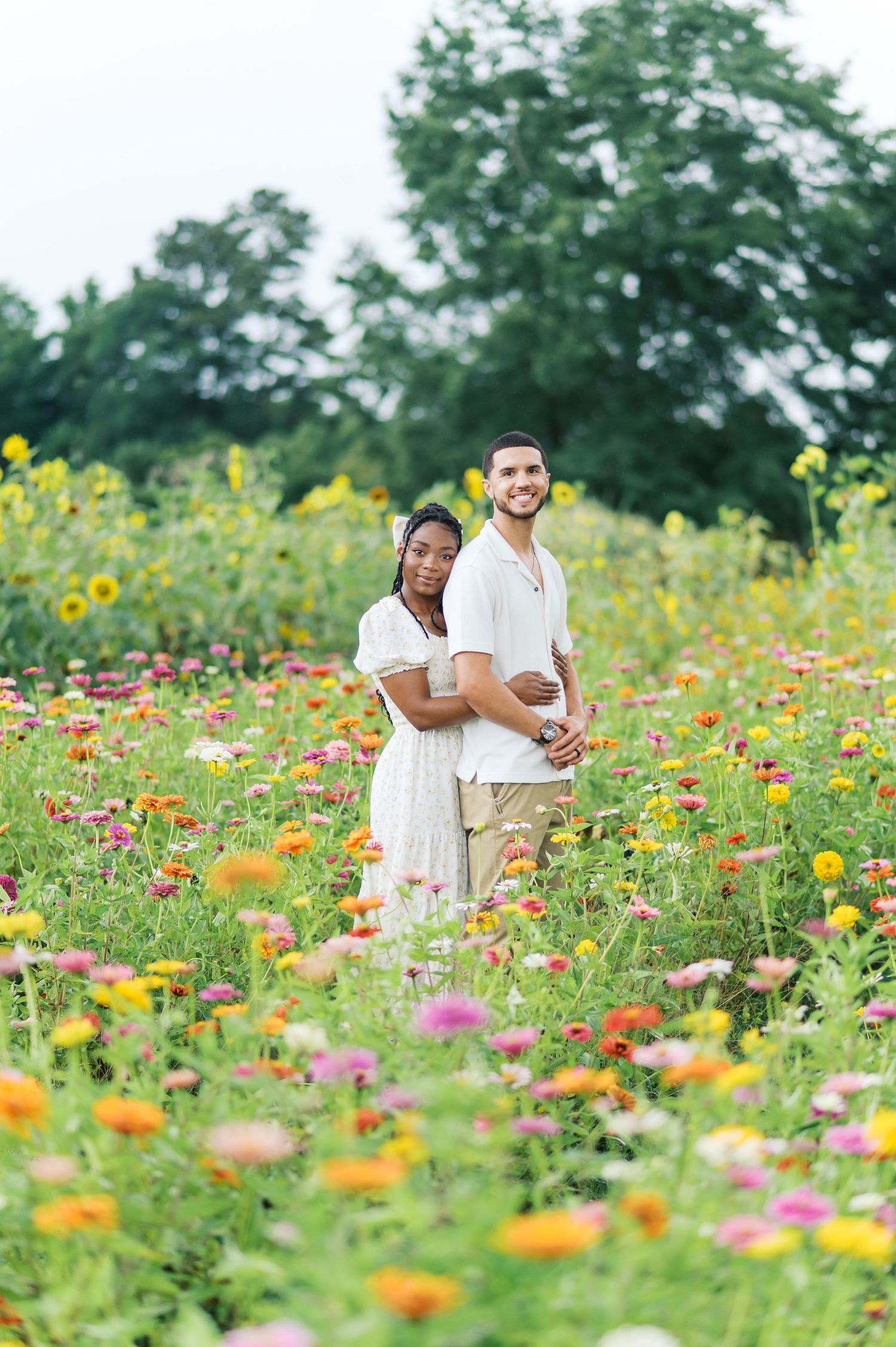 A couple embraces while standing in a vibrant wildflower field filled with zinnias, sunflowers and colorful summer blooms.