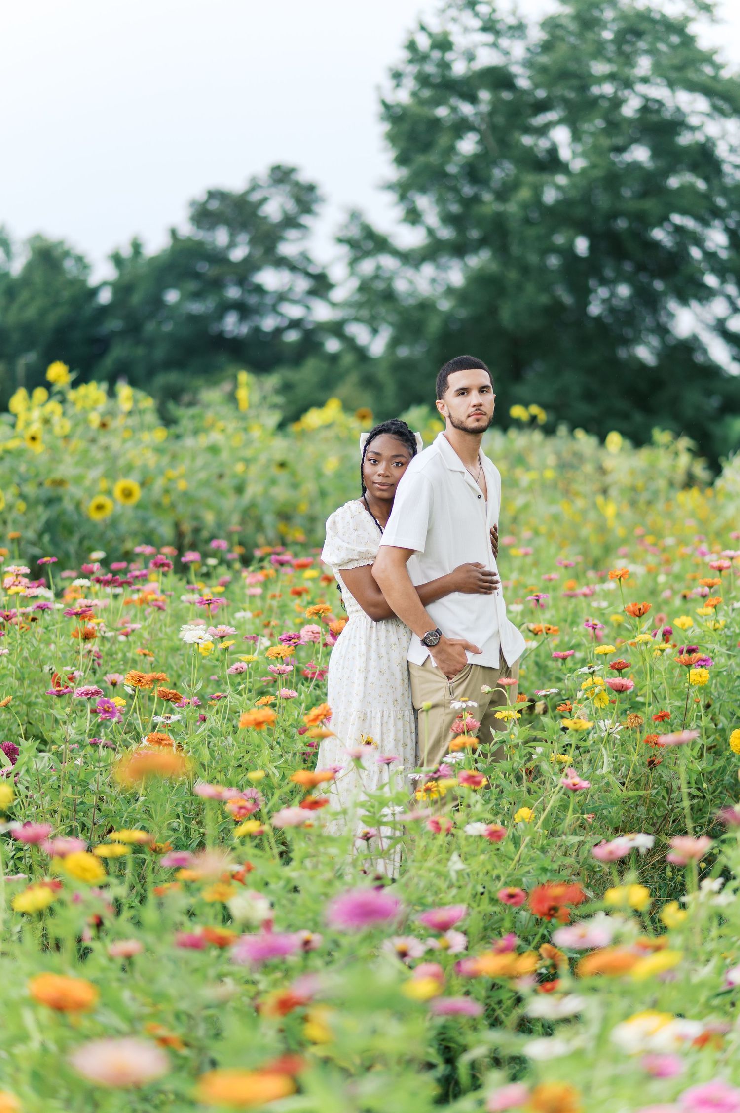 A couple embraces while standing in a vibrant wildflower field filled with colorful zinnias and sunflowers on a sunny day.