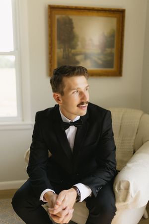 A person in a black tuxedo with a bow tie sits on a cream-colored couch beneath a landscape painting in a bright room.