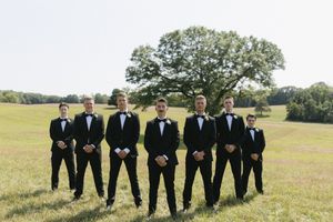 A group in matching black tuxedos and bow ties stand together in a line on a grassy field with trees in the background.