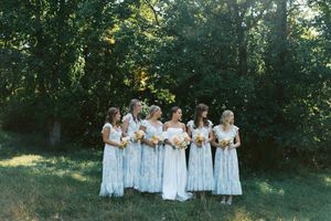 Bride and bridesmaids in floral blue dresses holding bouquets, standing together outdoors near lush green trees.