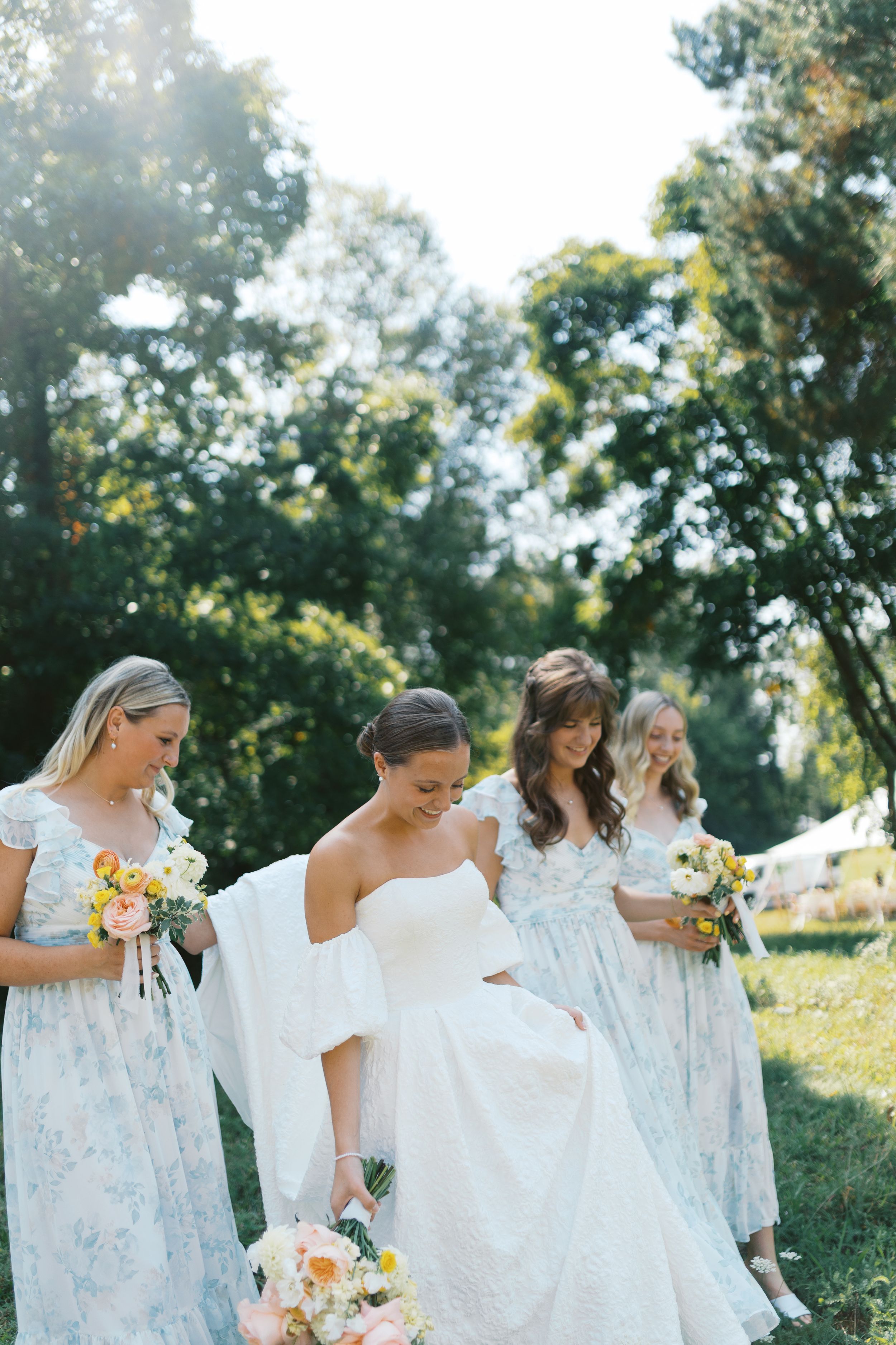Bride in white gown with bridesmaids in floral blue dresses holding colorful bouquets outdoors in a sunlit garden.