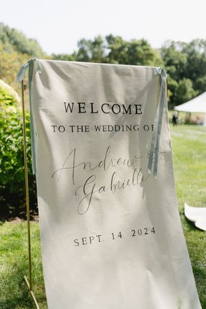 A beige wedding welcome sign with elegant calligraphy displaying the wedding date September 14, 2024 stands outdoors on grass.