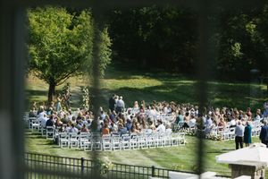 An outdoor wedding ceremony setup with white chairs arranged in rows on a sunny lawn surrounded by tall trees.