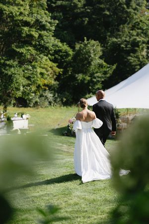 A couple in wedding attire walks toward a white tent on a lush green lawn surrounded by trees.