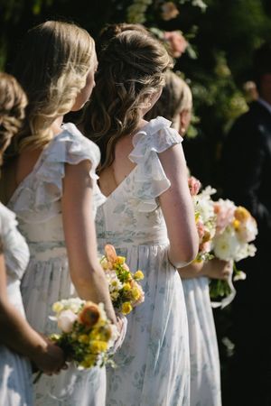 Bridesmaids in flowing white ruffled dresses hold yellow and white flower bouquets at an outdoor wedding.