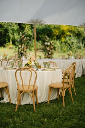 Elegant outdoor wedding reception table with white linens and wooden bentwood chairs set up on green lawn under tent.