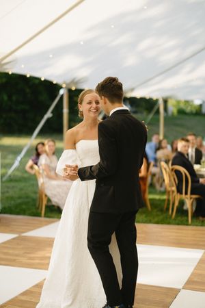 A couple shares their first dance on a checkered dance floor under an elegant outdoor wedding tent while guests look on.