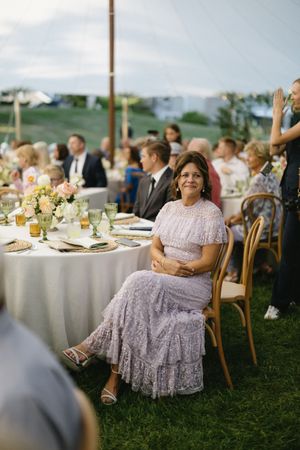 Outdoor wedding reception table set on grass with floral centerpieces and guests seated in wooden chairs.