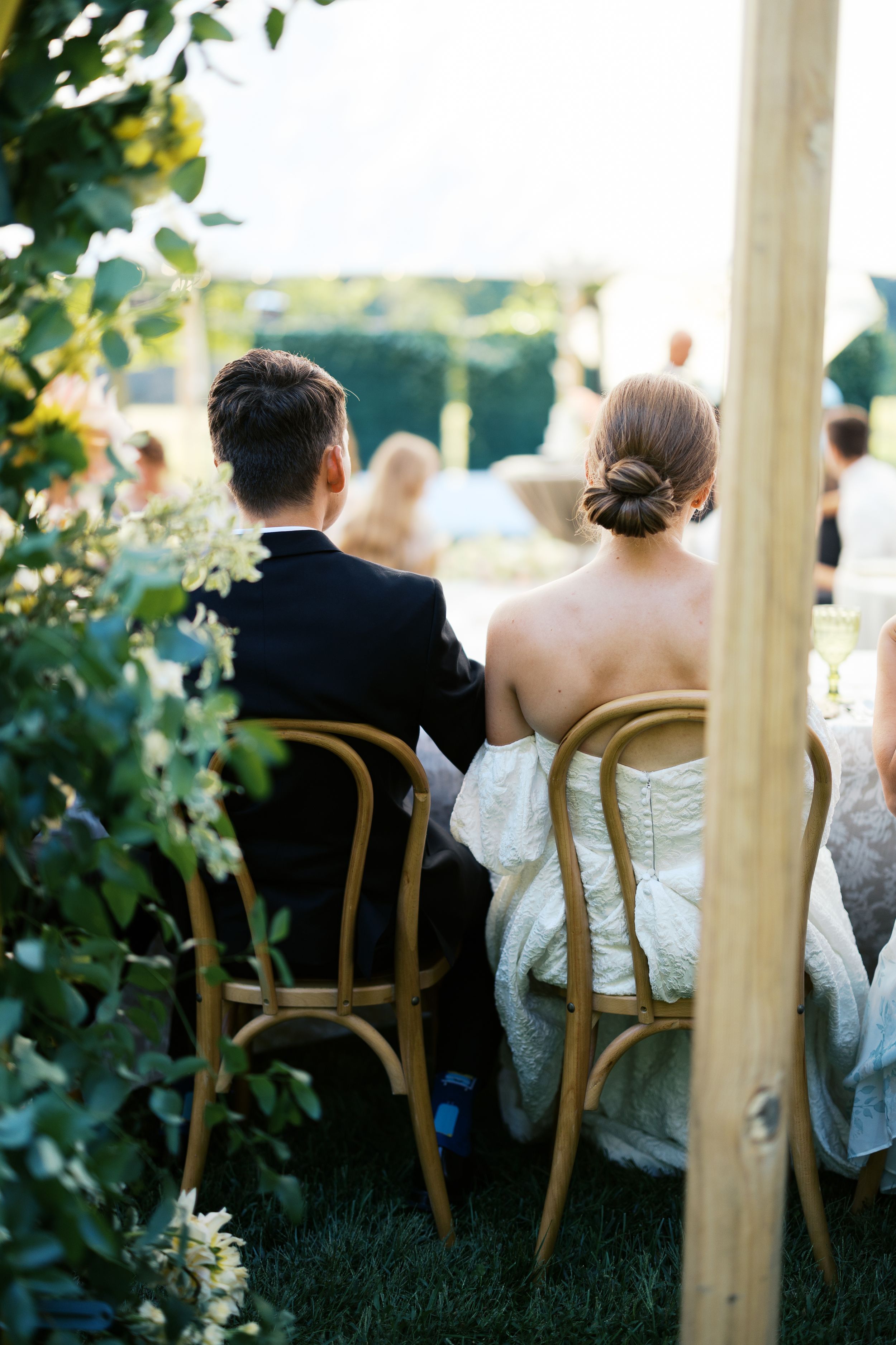 Bride and groom seated in wooden chairs at outdoor wedding ceremony, viewed from behind.