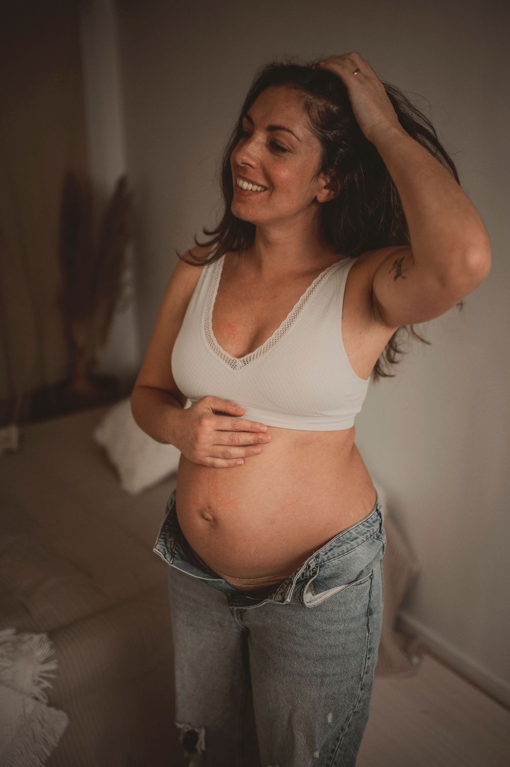 Pregnant woman in white bra top and jeans stands smiling with hand in hair in natural indoor lighting.