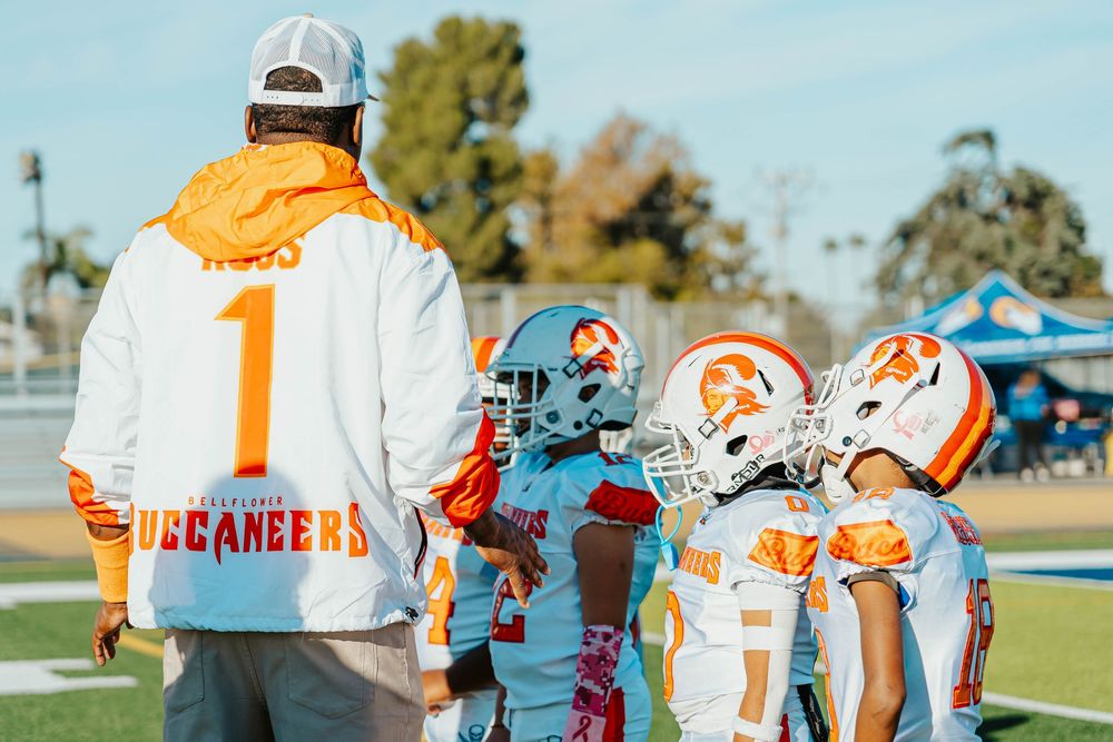 Baseball players in orange and white uniforms gather on a sunny field during a team practice session.