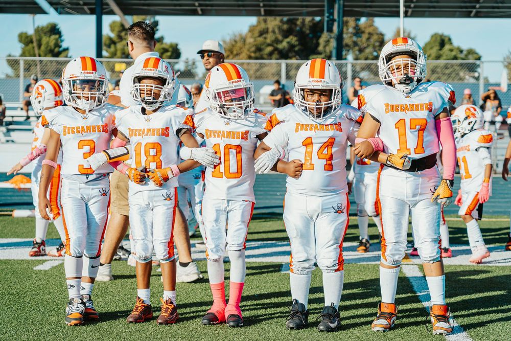 Football players in orange and white uniforms line up on a sunny field during practice or game preparation.