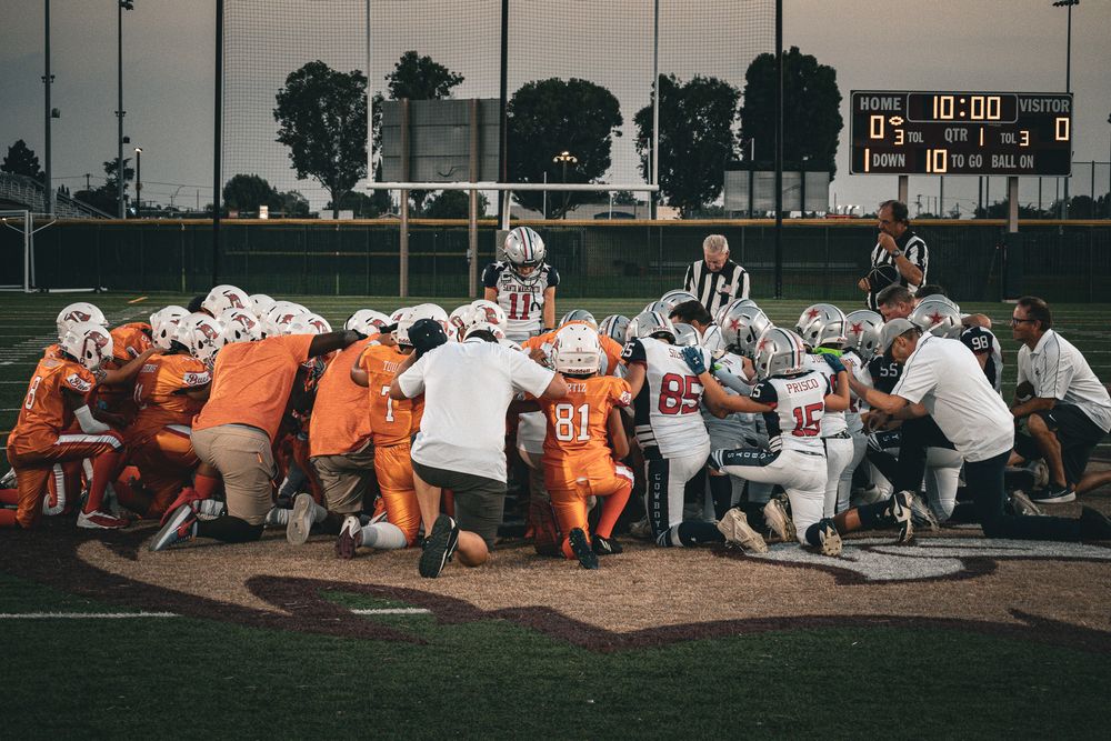 Football teams huddle together during a game, wearing orange and white uniforms on the field.
