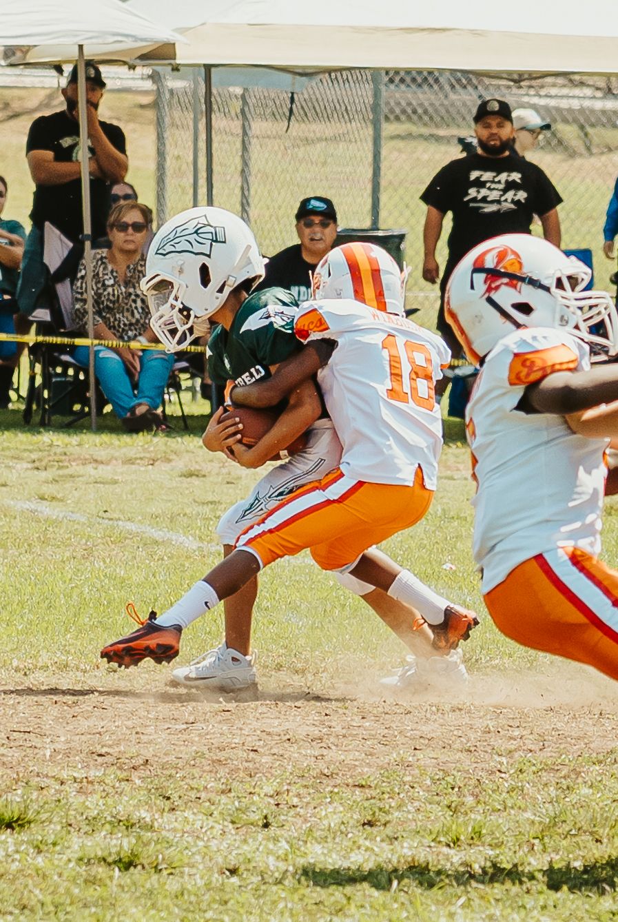 Youth football player in orange and white uniform carries the ball during a game on a grass field with spectators watching.
