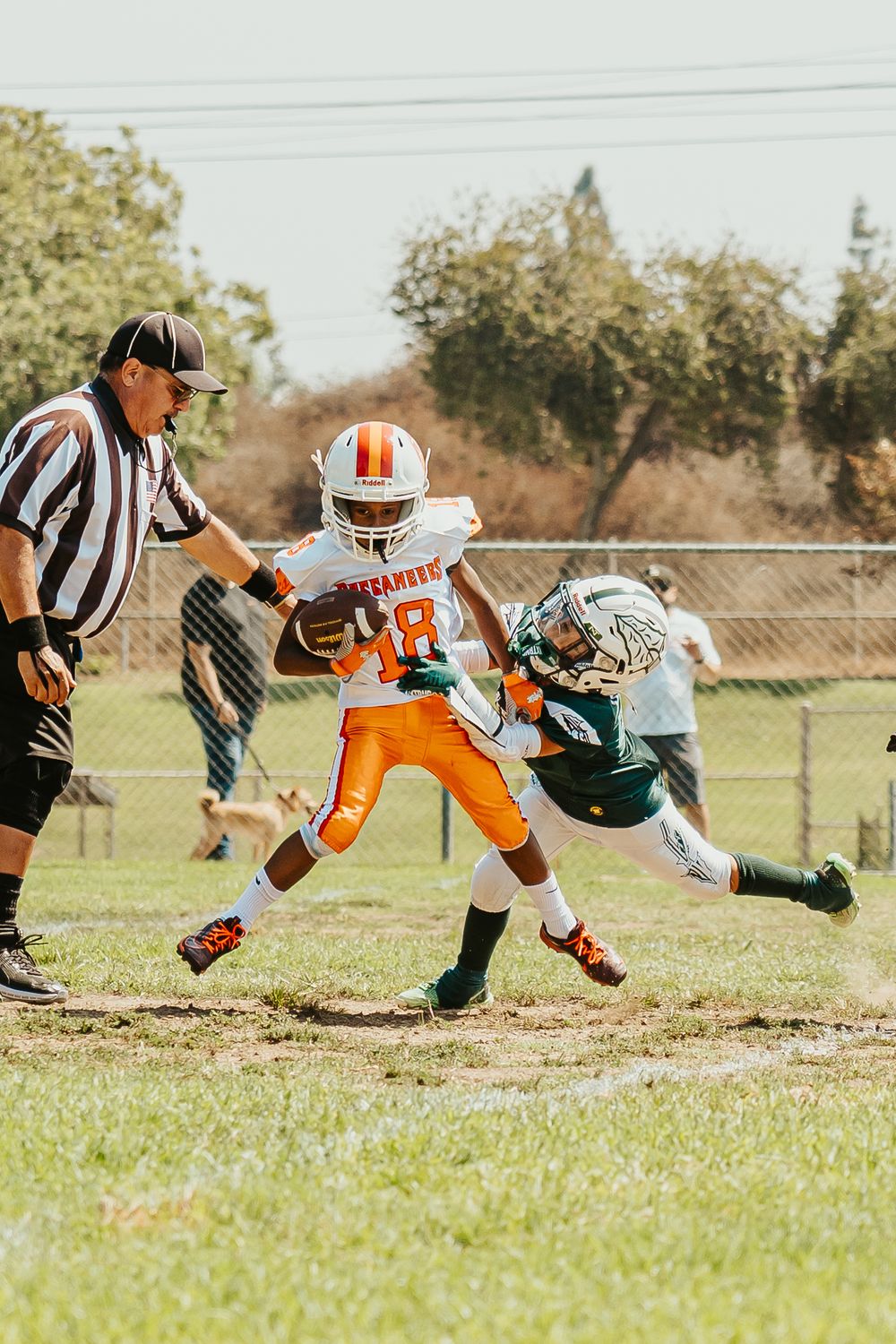 Youth football players in orange and green jerseys diving for a tackle during a game on a grass field.