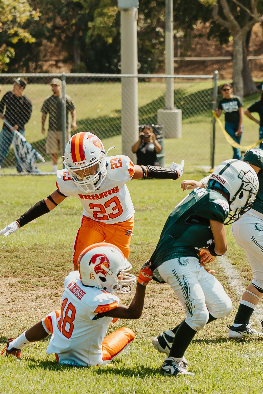 Football players in orange and white uniforms tackle an opponent in green during an outdoor game on a sunny day.