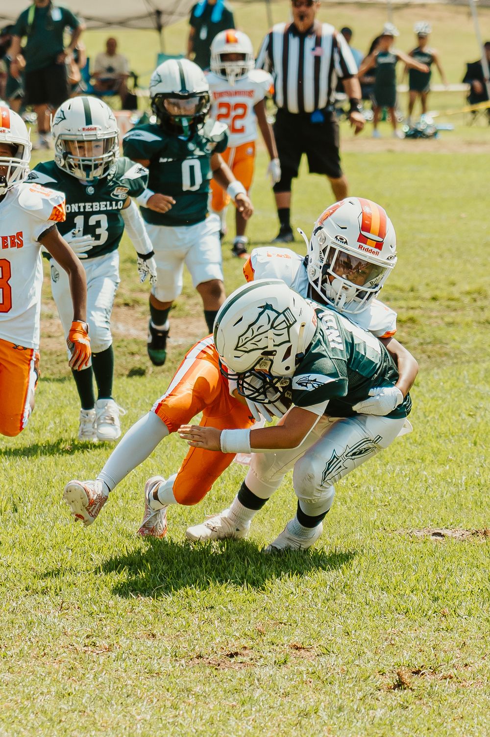 Youth football players in orange and green jerseys dive for a tackle during a game on a grass field.