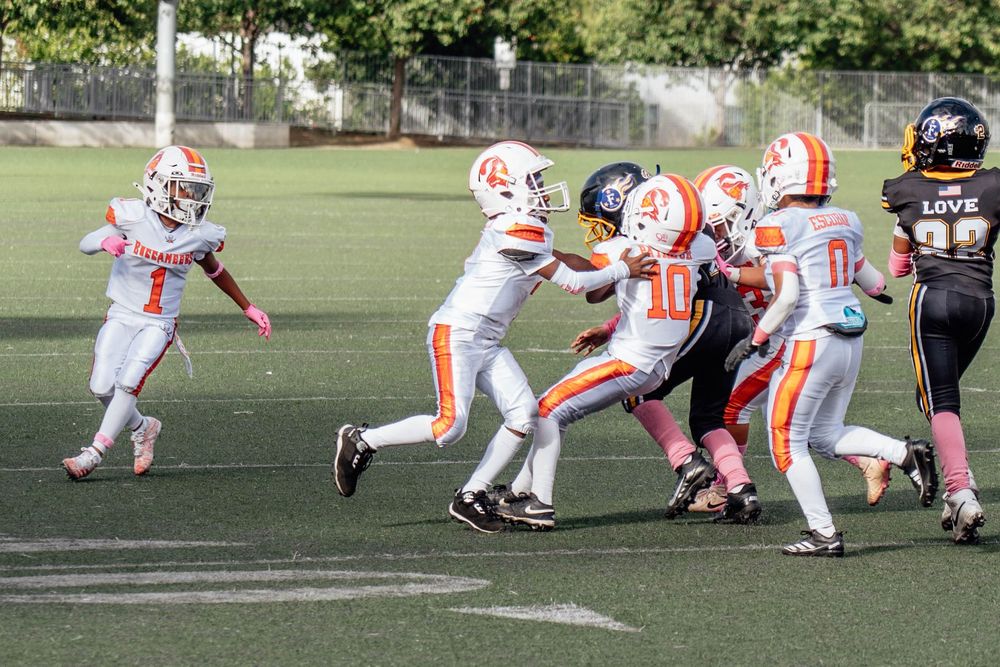 Youth football players in orange and white uniforms compete during a game on an outdoor turf field.