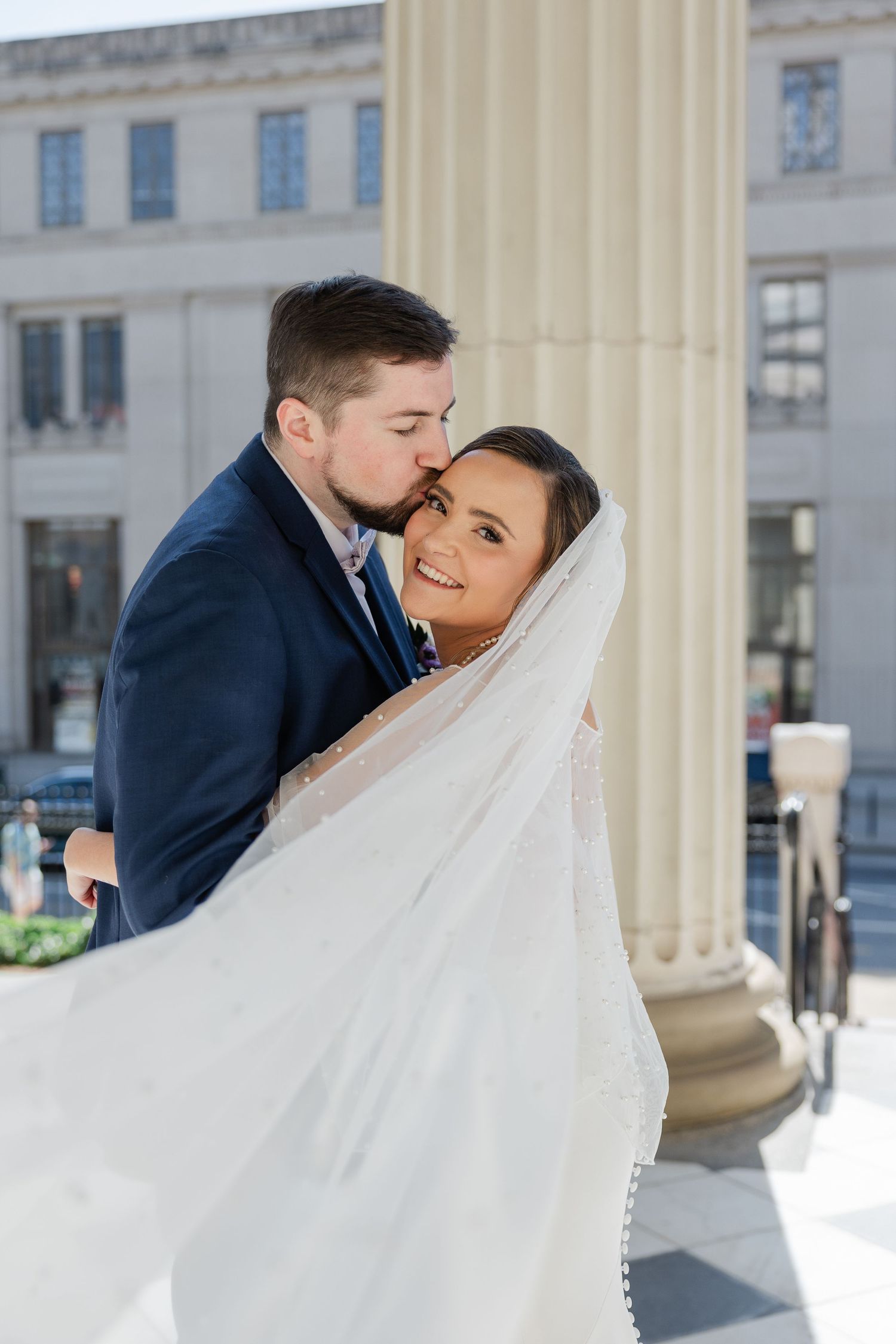 Intimate wedding portrait with flowing veil and white columns as couple shares tender embrace in navy and white attire.
