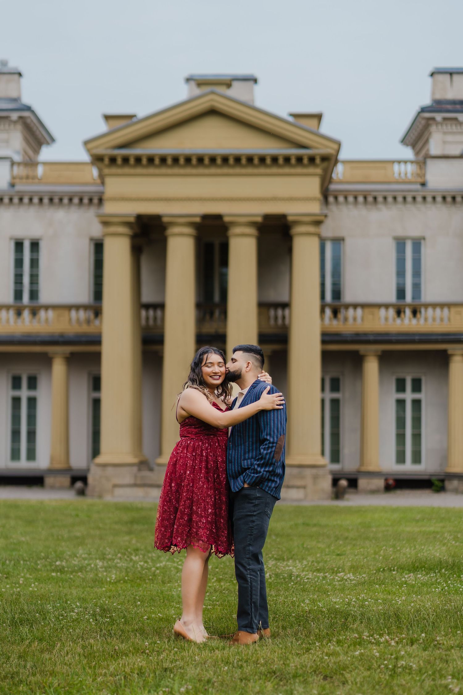 A romantic couple embraces on the lawn in front of a stately mansion with classical architecture and columns.