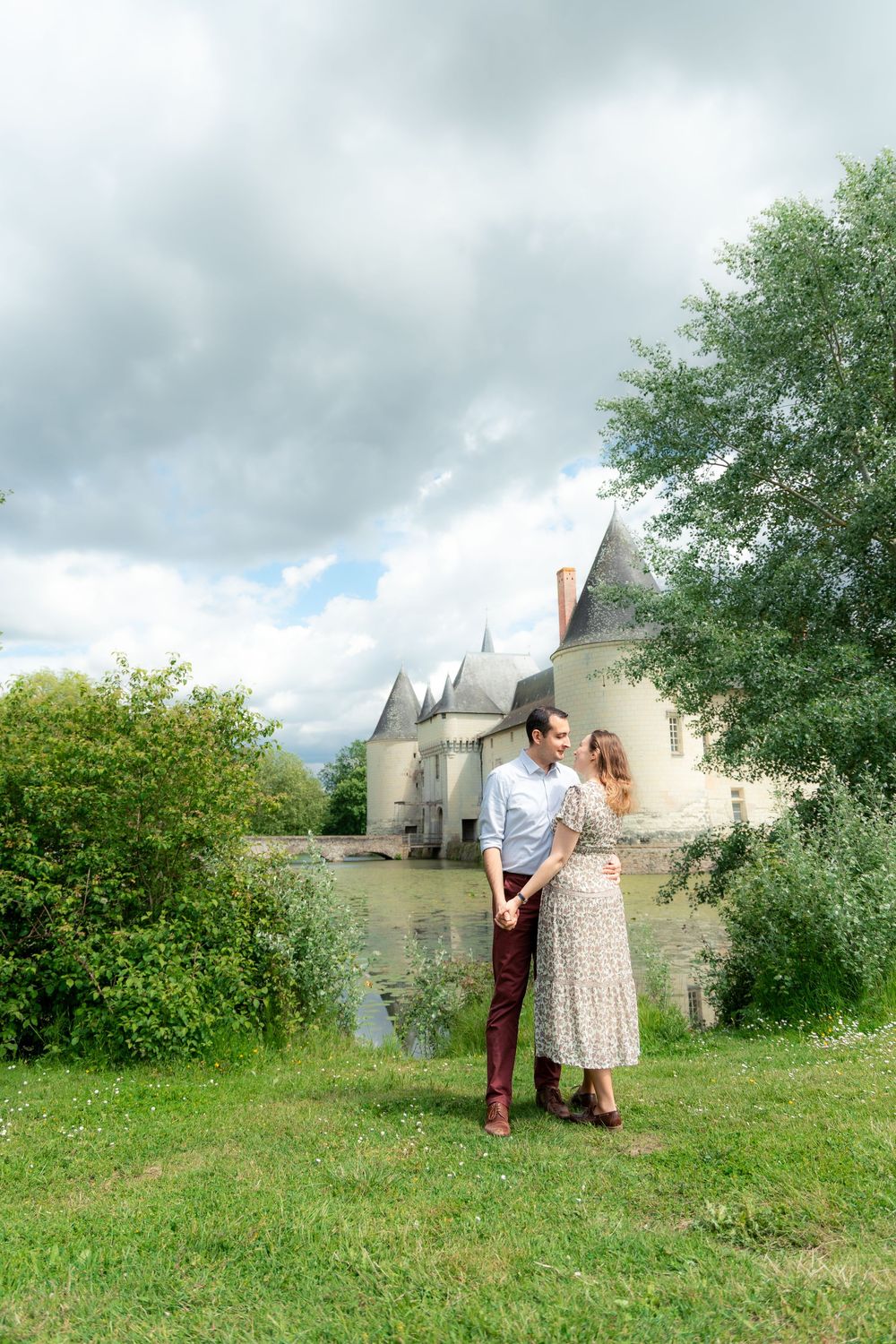 Couple holding hands on green lawn before a stunning French castle with moat and cloudy sky.