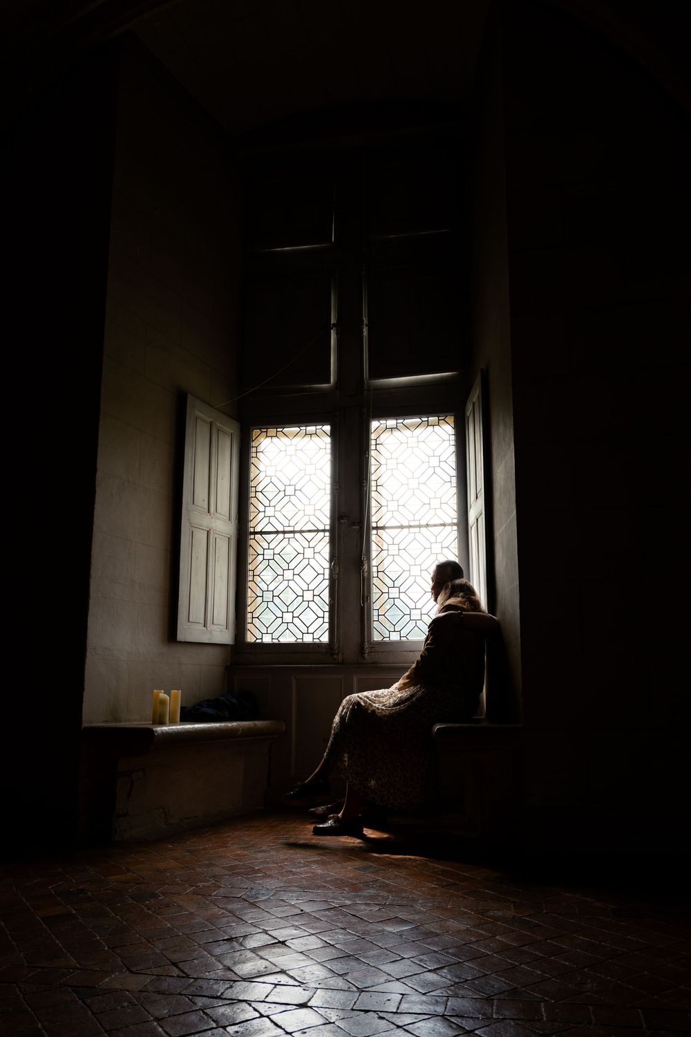 Solitary figure sitting by ornate window in dimly lit historic room with terracotta tile floor.