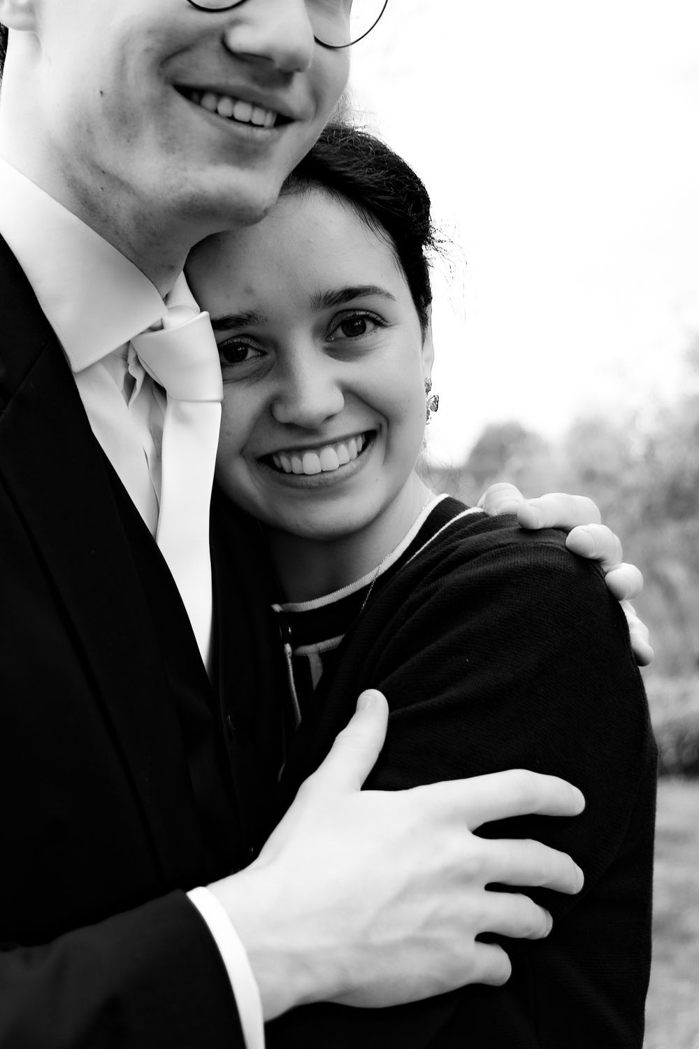 Black and white photo of a smiling couple embracing outdoors, man in suit hugging woman in dark jacket.