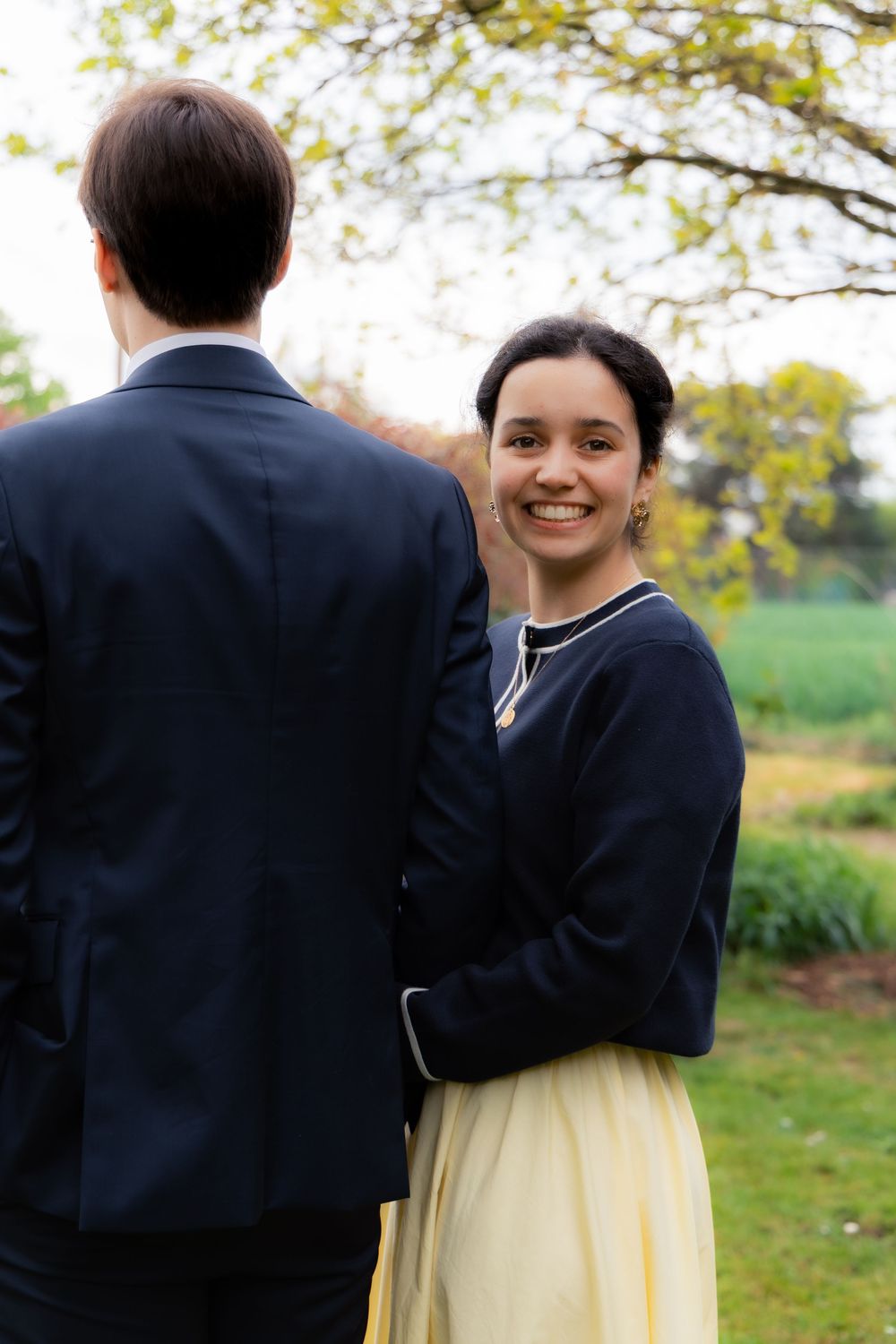 Smiling bride in navy jacket and yellow skirt glances back at groom in a beautiful garden setting.