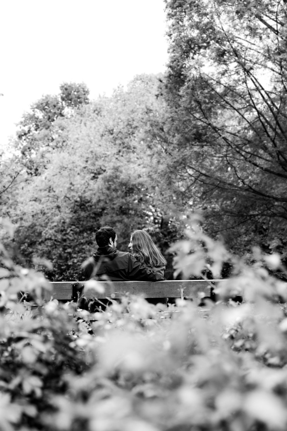 Black and white photo of a romantic couple sitting together on a bench surrounded by lush trees.