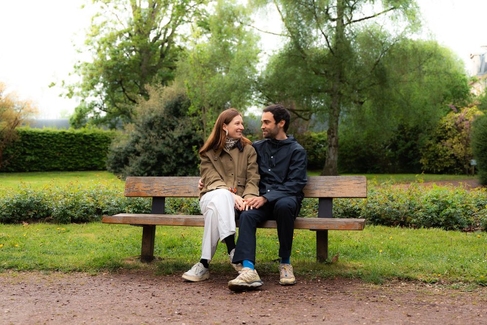 Couple sitting on a wooden park bench smiling at each other surrounded by lush green trees.
