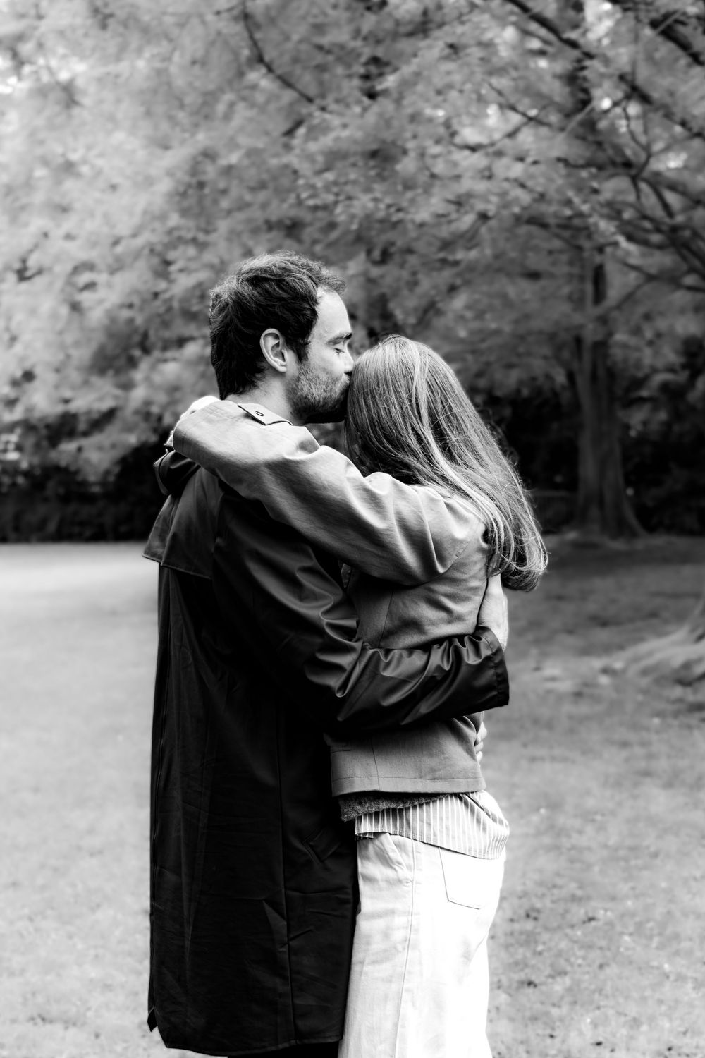 Black and white photo of a couple embracing and kissing in a park setting.