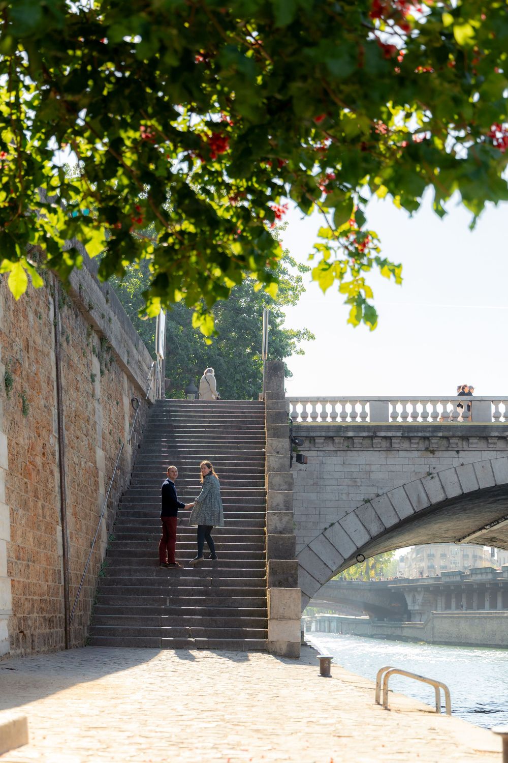 Couple holding hands on stone steps beneath a Parisian bridge framed by green foliage.