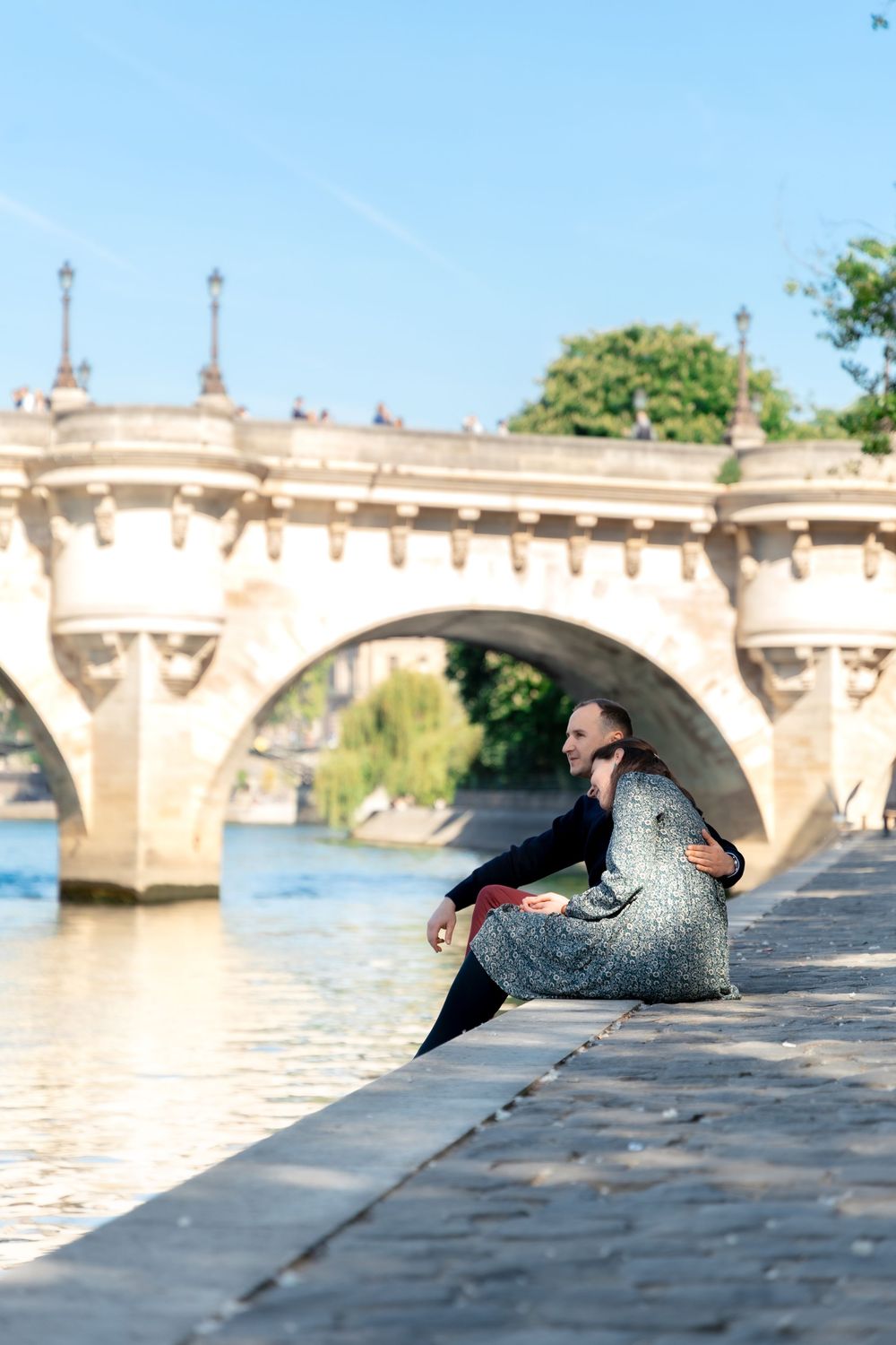 Woman in a floral dress sitting on stone steps by the Seine River with a bridge backdrop.
