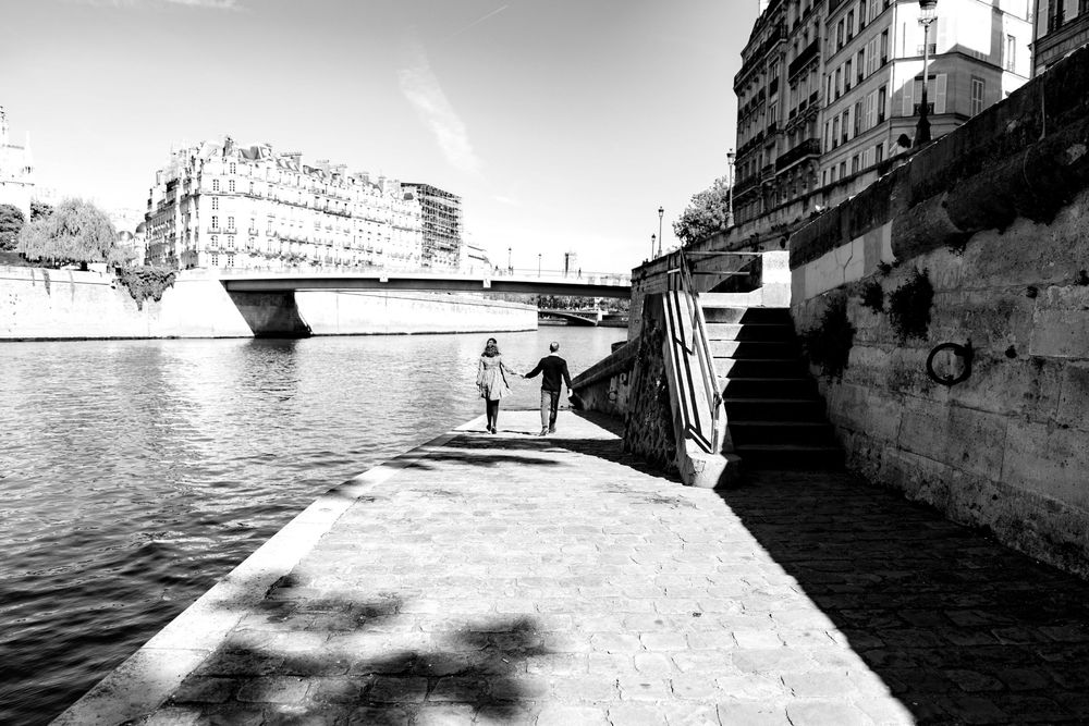 Black and white photo of a couple walking hand in hand along the Seine River in Paris.