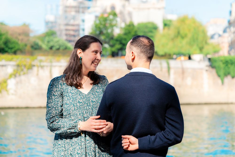Smiling couple holding hands by the Seine River in Paris during a romantic engagement moment.