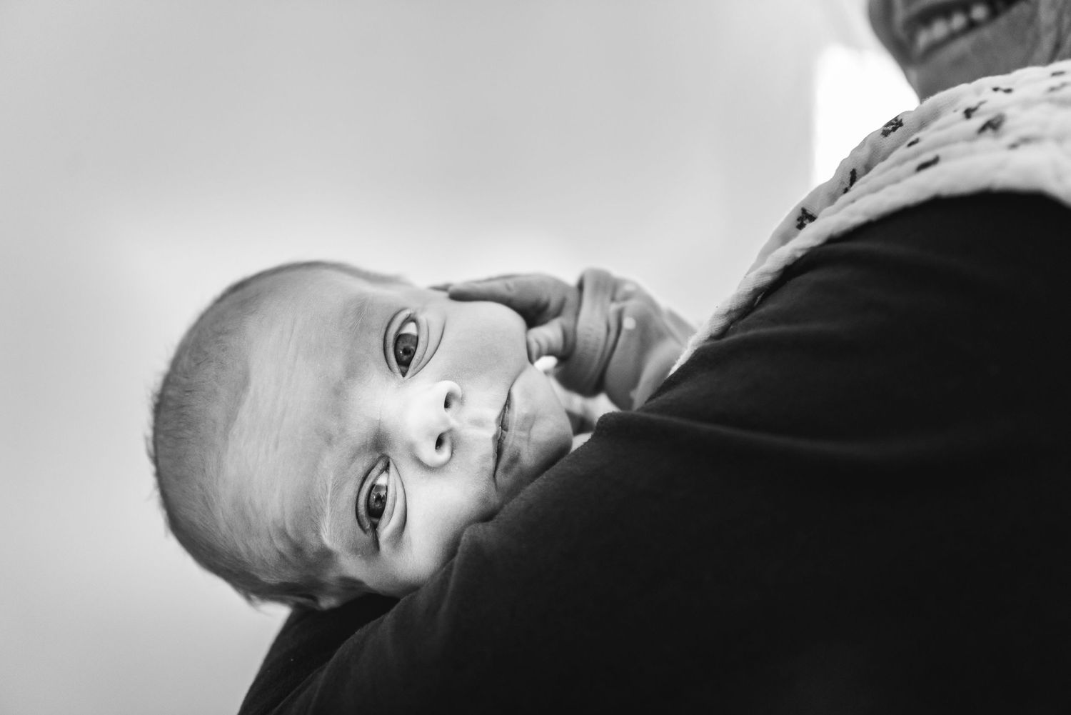 Close-up black and white shot of a baby looking up with curious eyes while being held against someone.