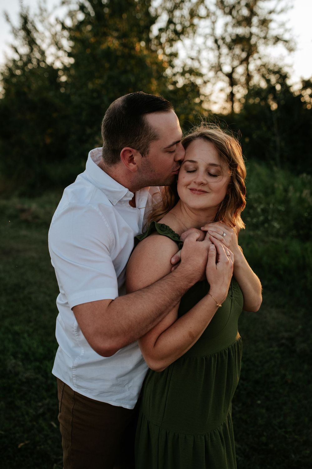 A romantic outdoor portrait of a couple embracing at sunset, with the woman in a green dress and man in white shirt.
