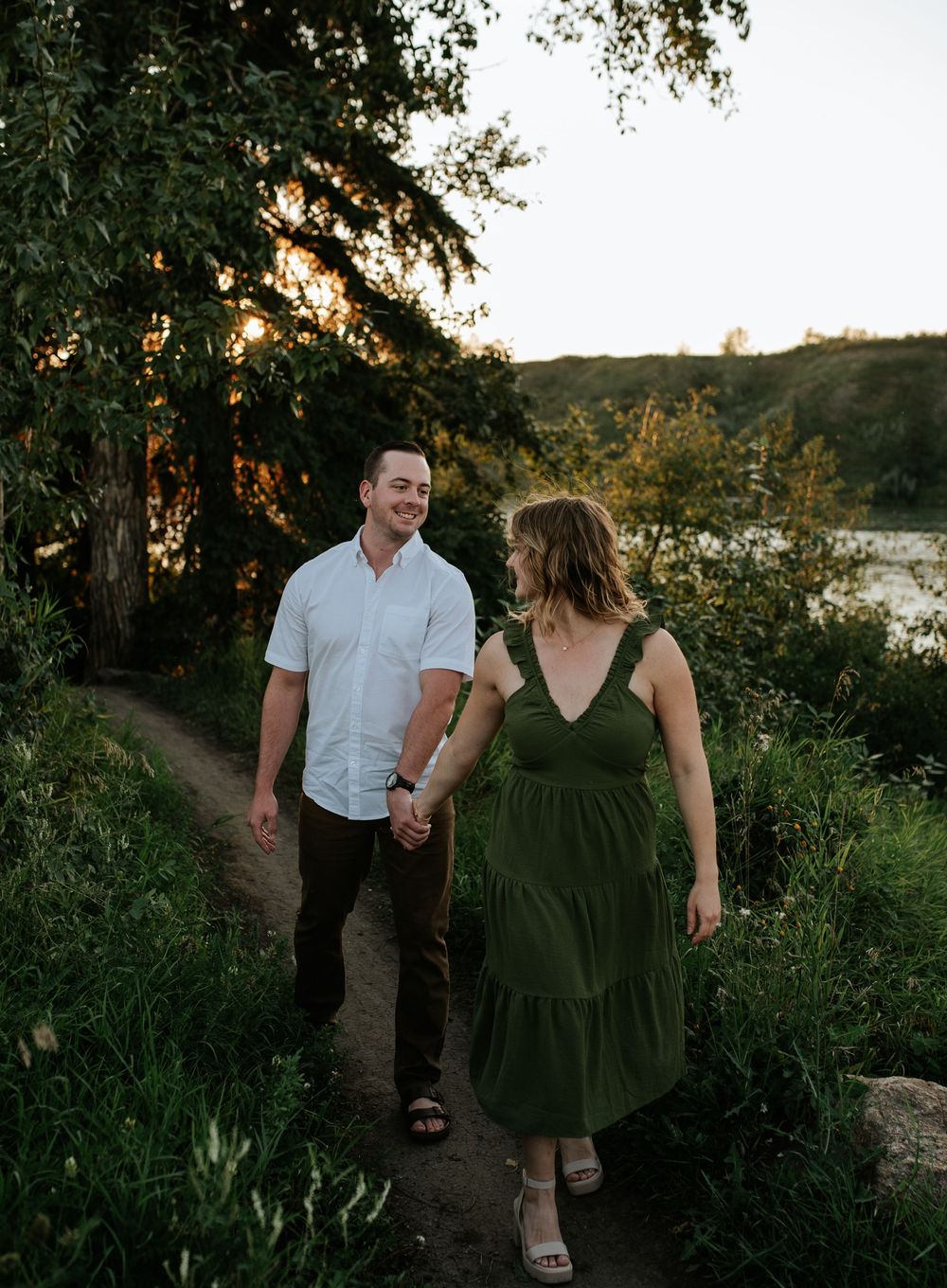 A couple holding hands walks along a scenic lakeside trail at sunset, surrounded by lush greenery and trees.