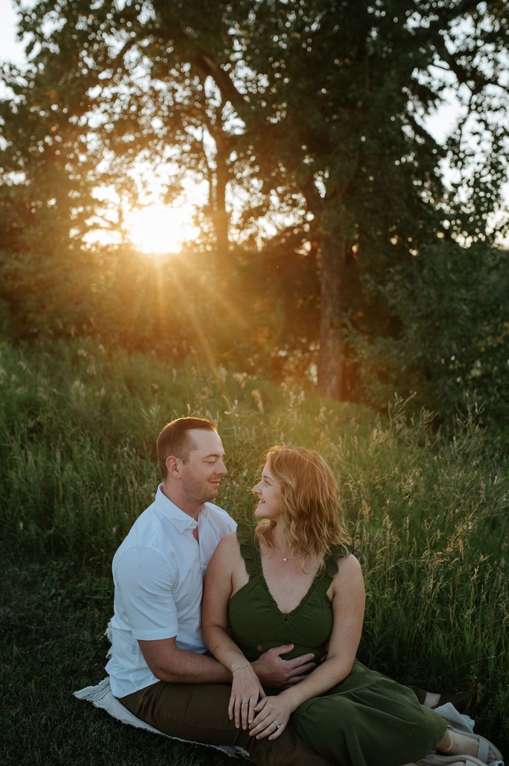 A romantic couple sits together in a grassy meadow during golden hour sunset with sun rays peeking through trees.