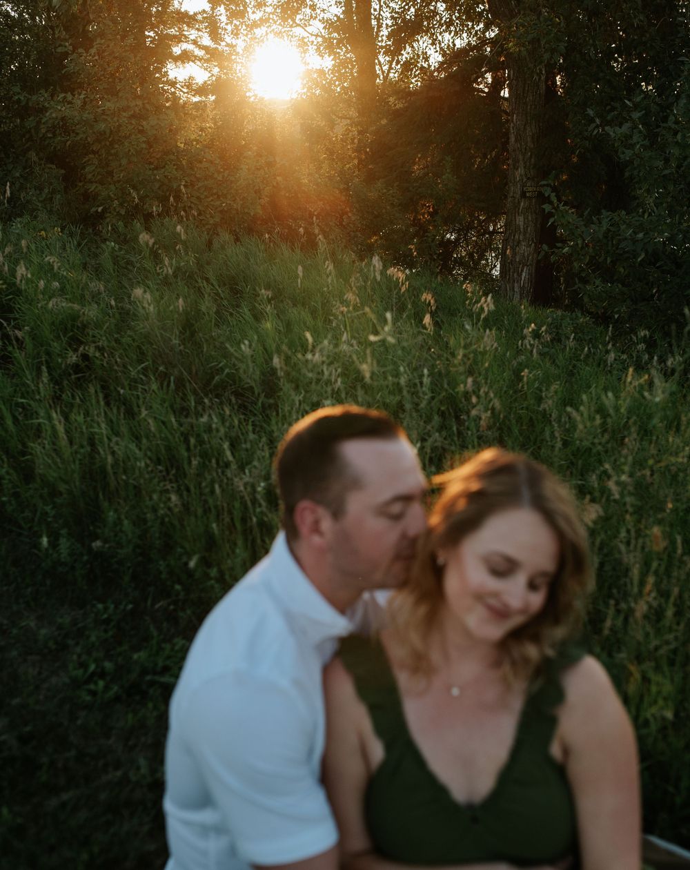 A couple shares an intimate moment at sunset with golden light filtering through trees in a natural outdoor setting.