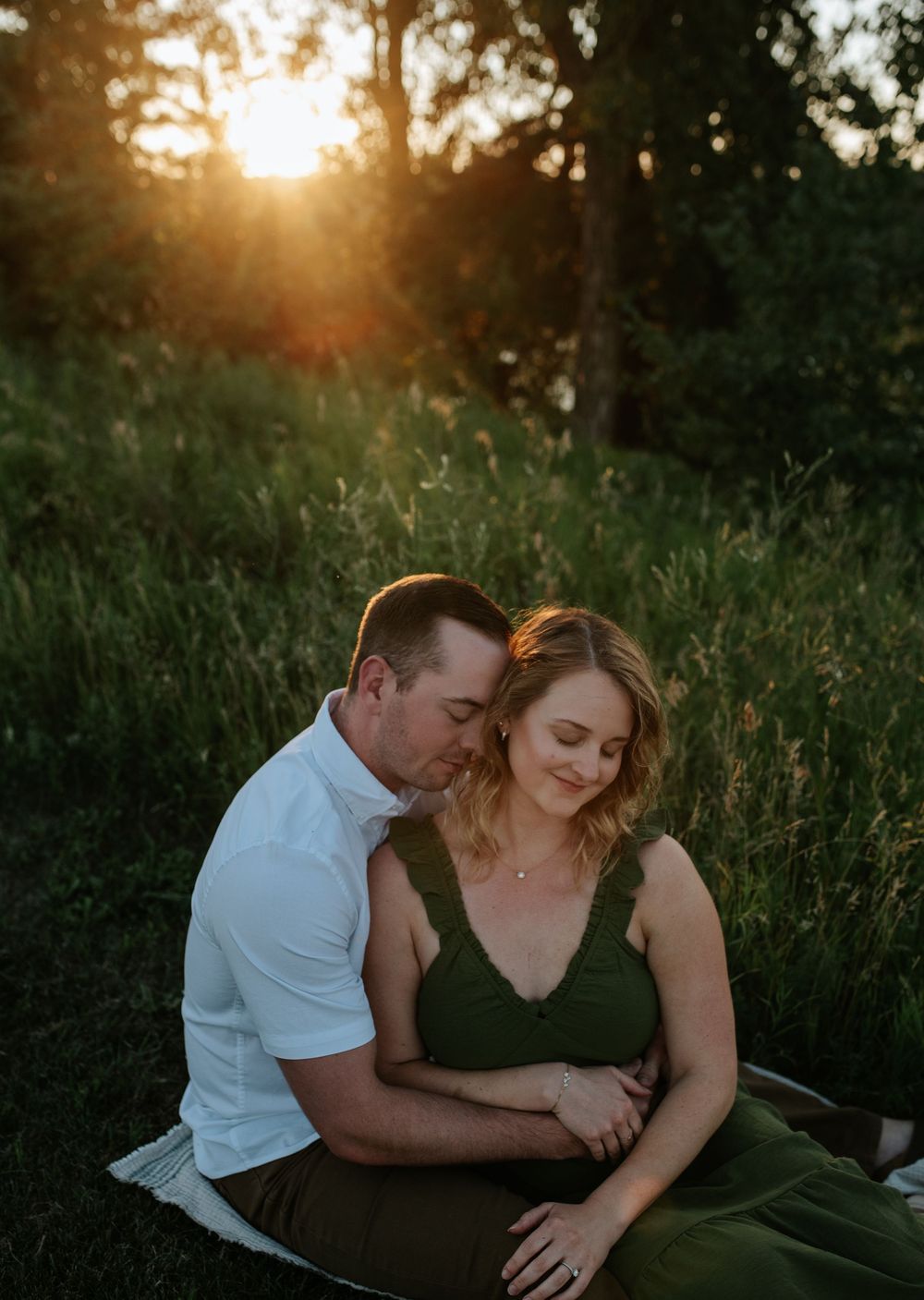 An expectant couple shares an intimate moment in a grassy field at sunset with beautiful golden backlighting.