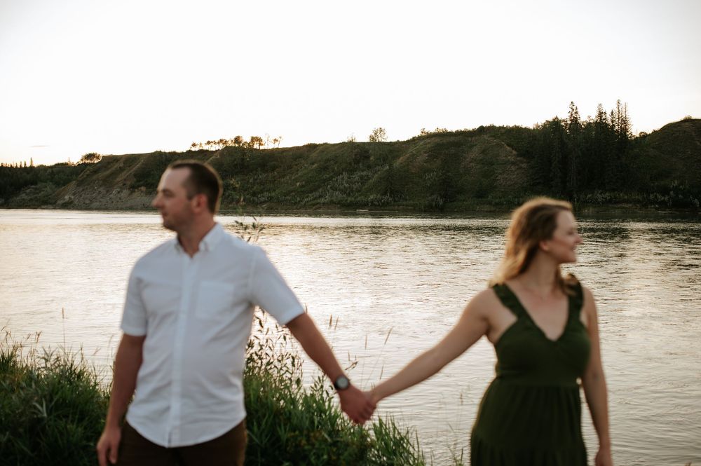 A couple holds hands while standing by a tranquil riverside at sunset with hills in the background.