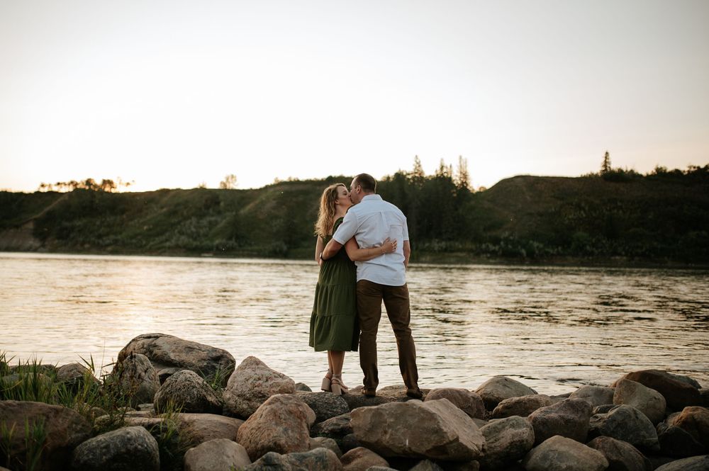 A romantic couple embraces on rocky riverbank during golden hour with cliffs in the distance.